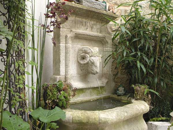 Fontaine de style ancien positionnée à l'angle d'un mur, avec tête de belier sculptée recrachant un jet d'eau régulier.