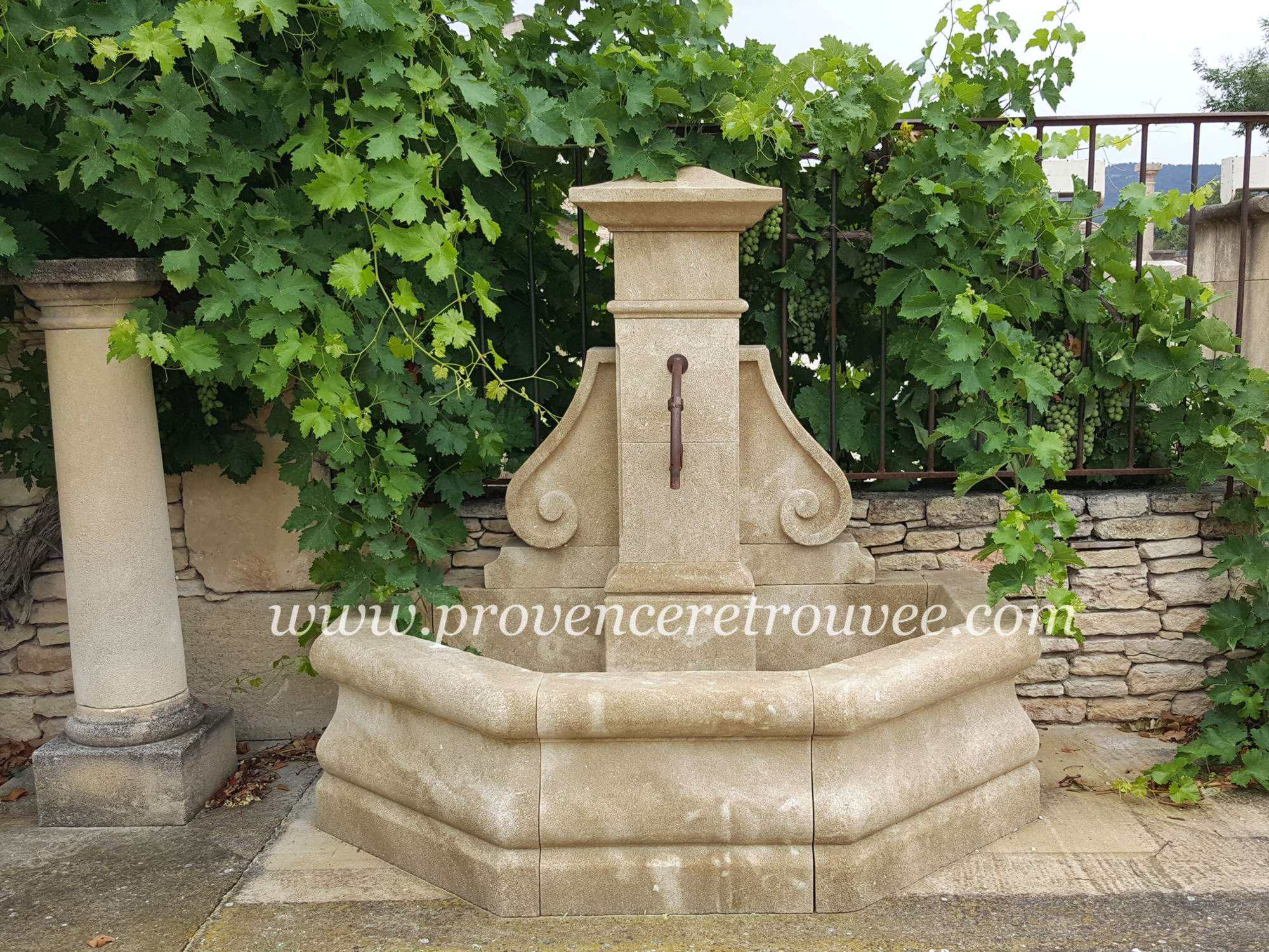 Fontaine extérieure en pierre taillée à la main Saint Tropez fon18-155 : Jolie fontaine murale d'exterieur disposant d'un bassin avec un grand volume d'eau. Le fronton se compose d'une colonne centrale habillée de part et d'autre de volutes sculptées.